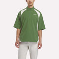 A man wearing the Reebok x Manors Men’s Tour Shirt by Reebok in green and white short-sleeves, paired with light-colored pants, stands against a plain white background.