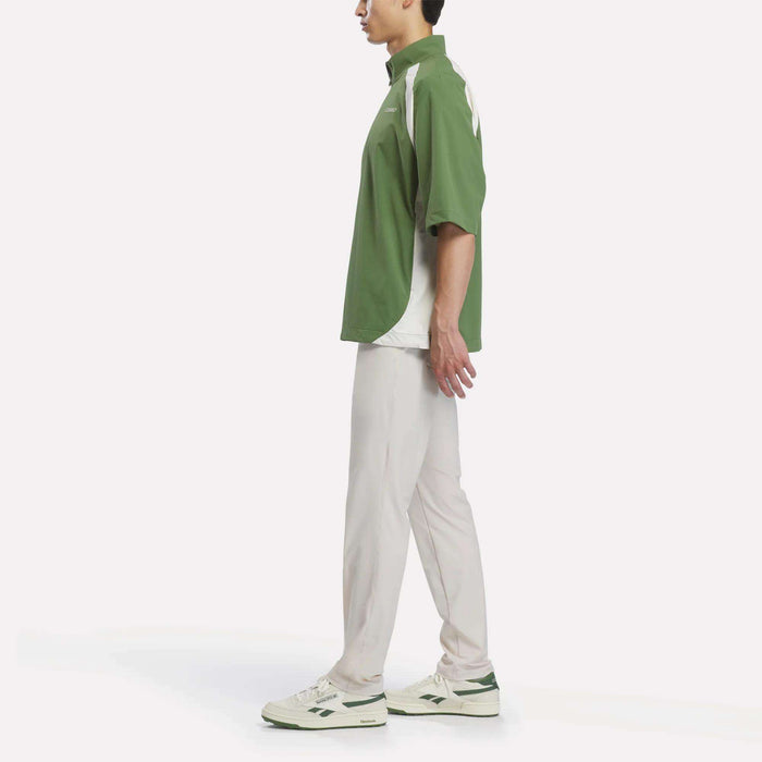 A person standing in profile, wearing a green and white Reebok x Manors Men’s Tour Shirt, light-colored pants, and white sneakers with green accents, against a plain white background.
