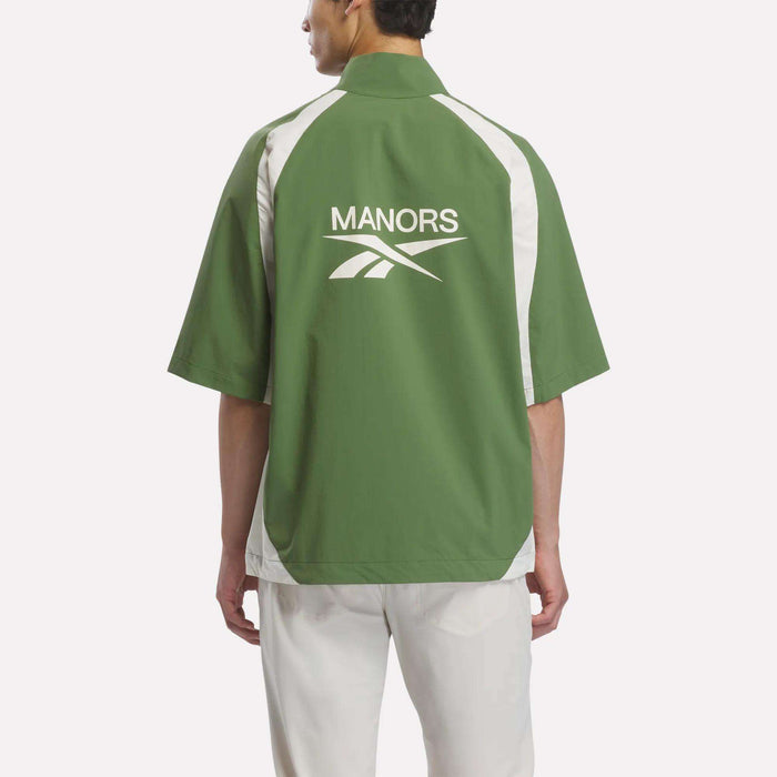 A person is shown from behind against a plain light background, wearing the Reebok x Manors Men’s Tour Shirt in green and white with short sleeves, featuring ‘MANORS’ and a logo on the back, paired with white pants.