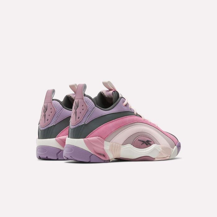The Reebok Shaqnosis Low Shoes feature a pink, purple, and mauve layered design with Hexalite cushioning for comfort. Brand logos appear on the side and heel. The shoes are angled left on a white background.