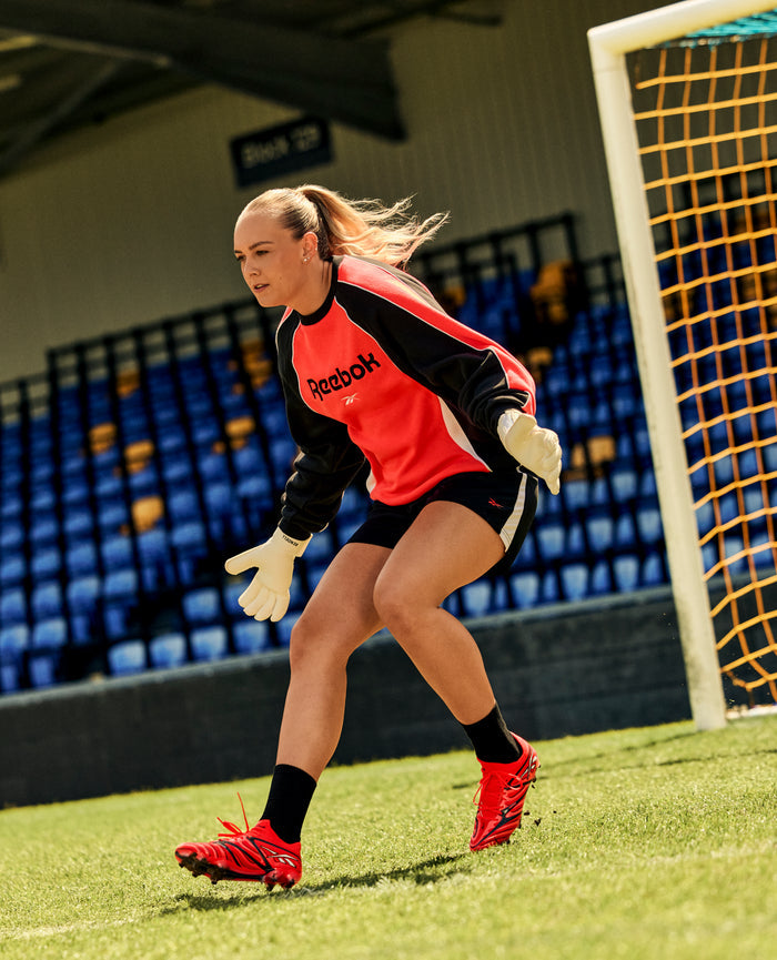 A female soccer goalkeeper in a red Reebok jersey and gloves stands focused in front of the goal on a sunny day, wearing Reebok Sidewinder 26 Elite Firm Ground Cleats planted firmly on the well-maintained grass, with empty blue and yellow stadium seats in the background.