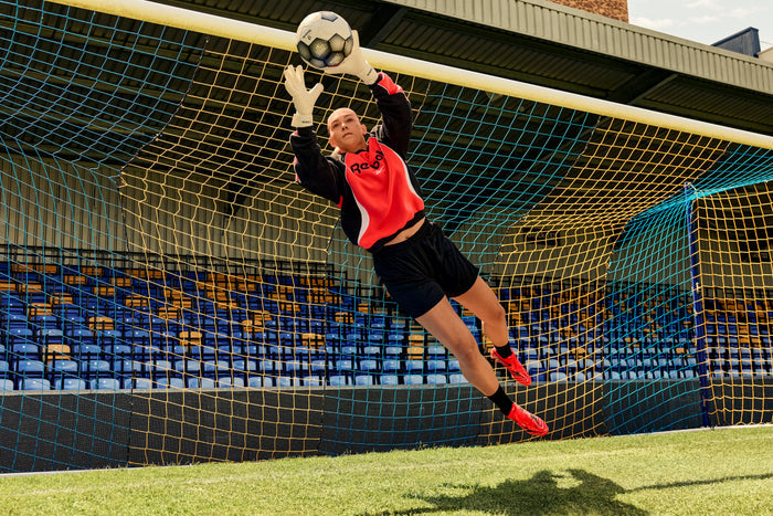 A soccer goalkeeper in a red and black jersey, wearing Reebok Sidewinder 26 Elite Firm Ground Cleats, leaps to catch a ball mid-air in front of the goal during daylight, with empty stadium seats visible in the background.