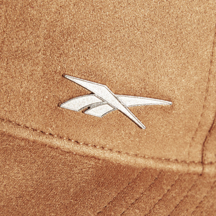 A close-up of the brown Suede Badge Cap by Reebok shows a silver, angular metallic logo, giving this vegan suede cap a modern look.