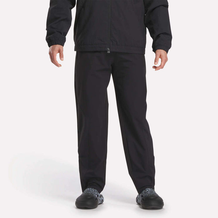 A person is shown from shoulders down wearing Reebok Track Pants, a black jacket, and black shoes against a plain white background.