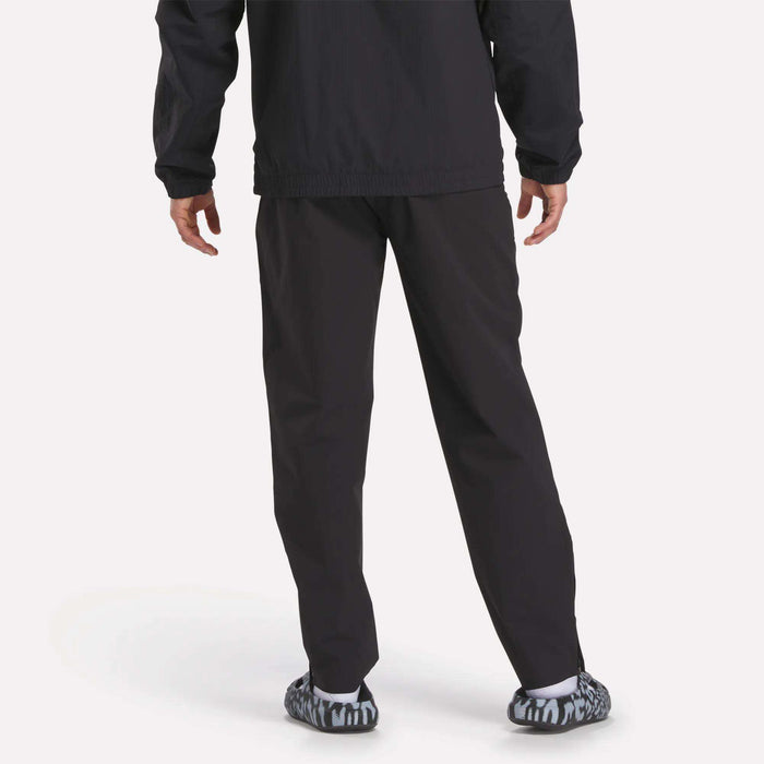 A person stands facing away in Reebok Track Pants, paired with a black jacket and black-and-white patterned slip-on shoes, against a plain light background.