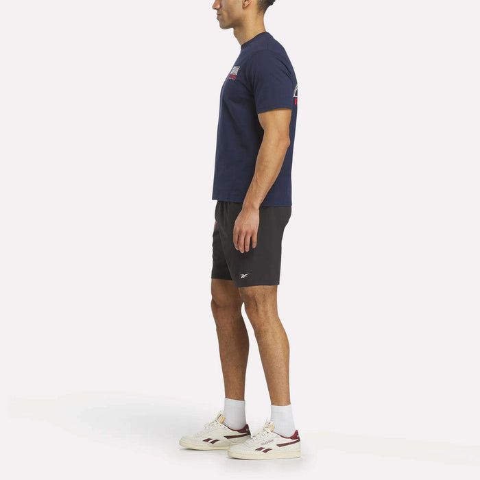 A man in a navy blue Reebok USA Football T-Shirt, black shorts, and white sneakers stands sideways against a plain white background.