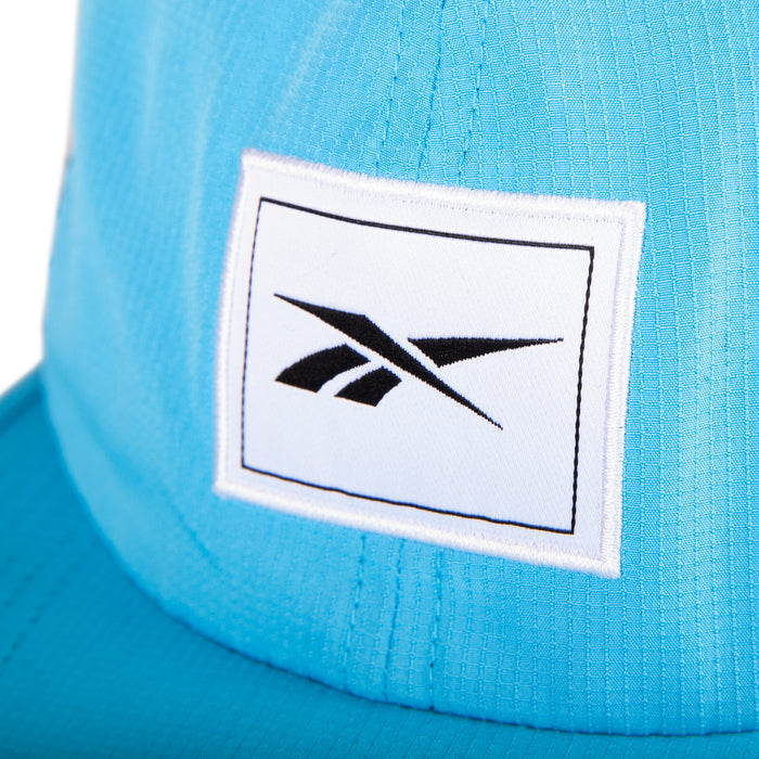 Close-up of the Reebok Vented Flat Brim Running Cap in blue, showcasing a white rectangular patch with a black Reebok logo on the front.