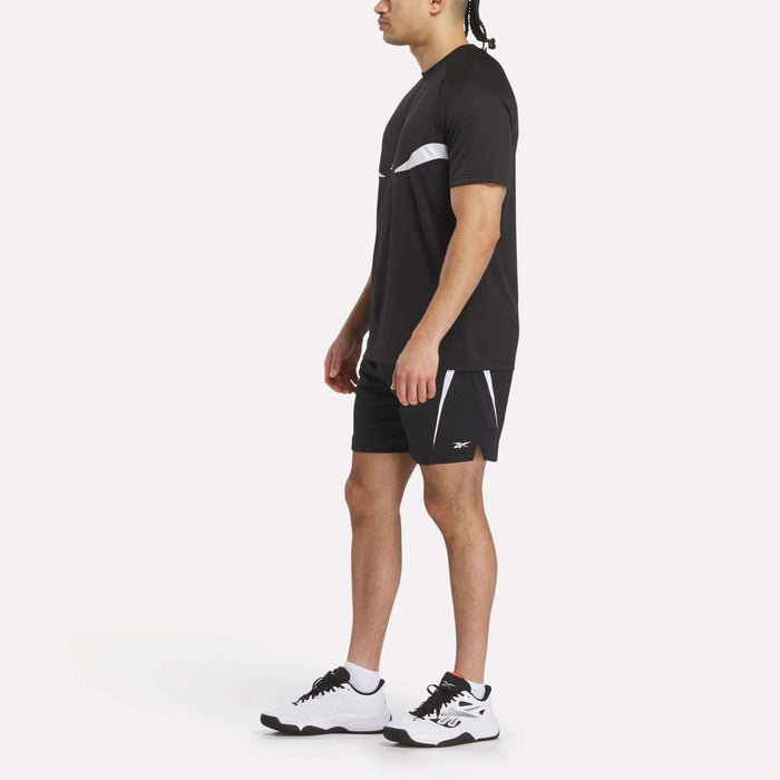 A man stands in profile on a plain white background wearing a black Reebok Warm-Up Basketball T-Shirt, black athletic shorts with white accents, and white sneakers.