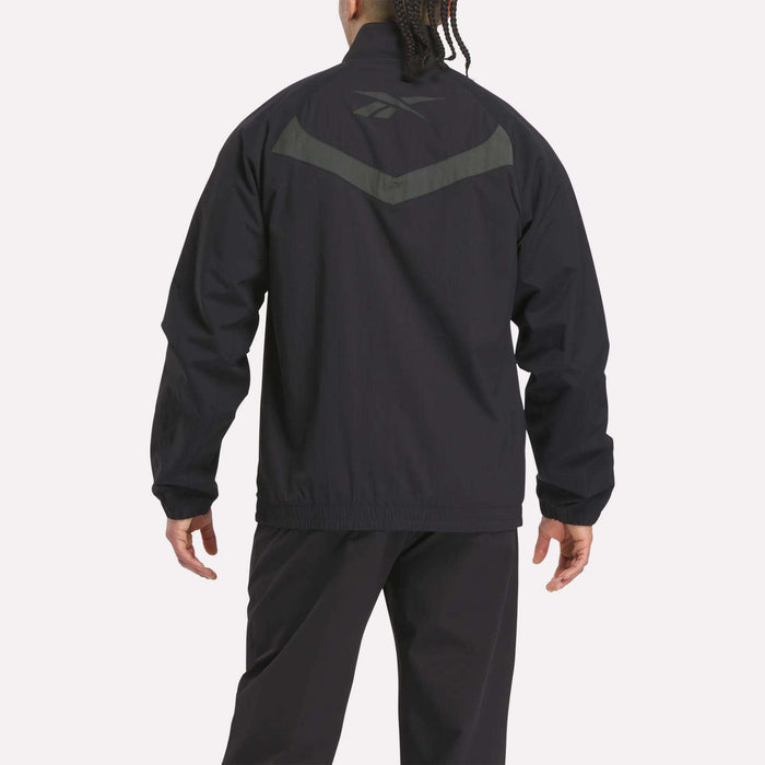 A person faces away, wearing a black Reebok Warm Up Track Jacket and pants. The jacket has a dark gray Reebok logo and chevron on the upper back. The background is plain and light-colored.