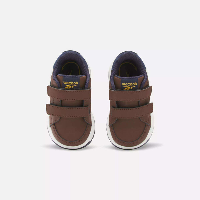 The Reebok Weebok Clasp Low Shoes for infants and toddlers are brown with white soles, double Velcro straps, a dark blue inner lining, and yellow tongue accents, shown facing forward on a plain light background.