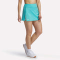 A person in a turquoise Reebok Women's Athlete Speed Skort 3" with built-in shorts, ribbed waistband, and small logo near the hem pairs it with white sneakers against a plain white background.