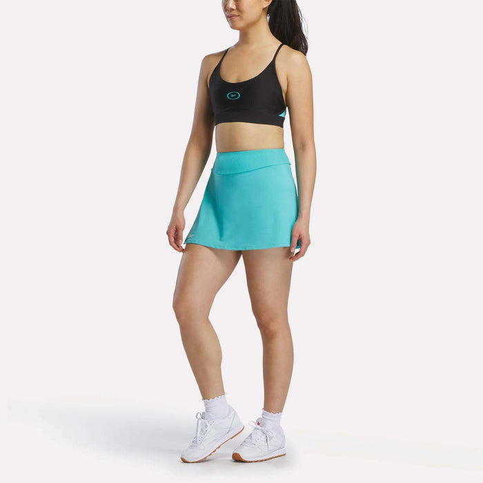 A woman in a black sports bra, Reebok Women's Athlete Speed Skort 3" in turquoise, white socks, and white sneakers stands against a plain white background.