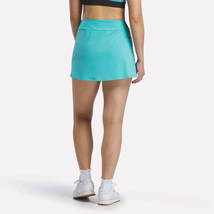 A woman stands with her back to the camera, wearing a turquoise Reebok Women's Athlete Speed Skort 3" with a ribbed waistband, a black and turquoise sports top, white socks, and white sneakers against a plain white background.