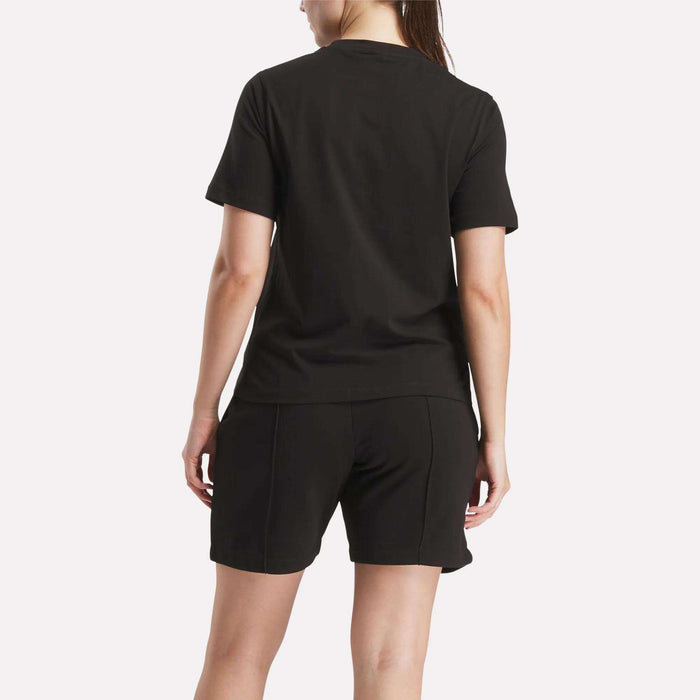A person faces away from the camera, wearing a Reebok Women's Big Logo Crewneck Short Sleeve Tee in black and black shorts against a light background.