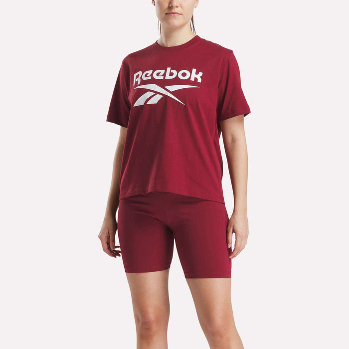 A person is wearing Reebok's Women's Big Logo Oversized Crew Short Sleeve Tee in maroon with a large white logo, paired with matching maroon bike shorts, standing against a plain white background.