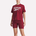 A person is wearing Reebok's Women's Big Logo Oversized Crew Short Sleeve Tee in maroon with a large white logo, paired with matching maroon bike shorts, standing against a plain white background.