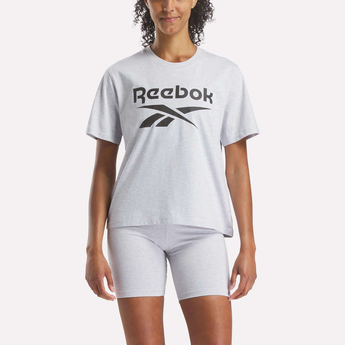 A person stands against a white background wearing a Reebok Women's Big Logo Oversized Crew Short Sleeve Tee in light gray with a black logo, paired with matching gray shorts.