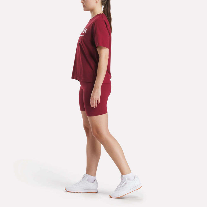 A woman in profile wears a Reebok Women's Big Logo Oversized Crew Short Sleeve Tee in burgundy, matching bike shorts, and white sneakers, her hair tied back, standing against a plain white background.