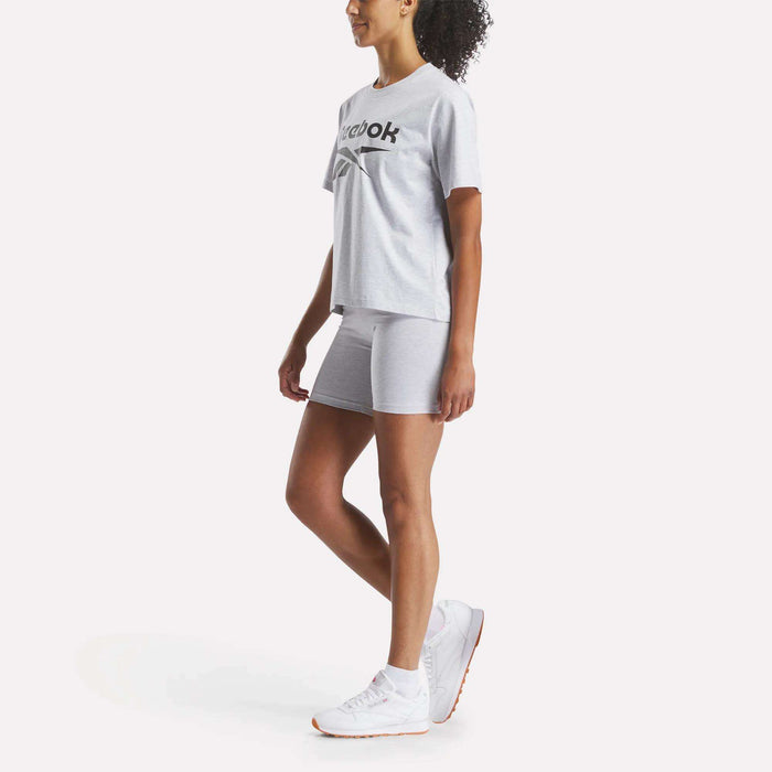 A woman stands in a Reebok Women's Big Logo Oversized Crew Short Sleeve Tee, paired with matching shorts and white sneakers, set against a plain white background. She is viewed from the side and looking forward.