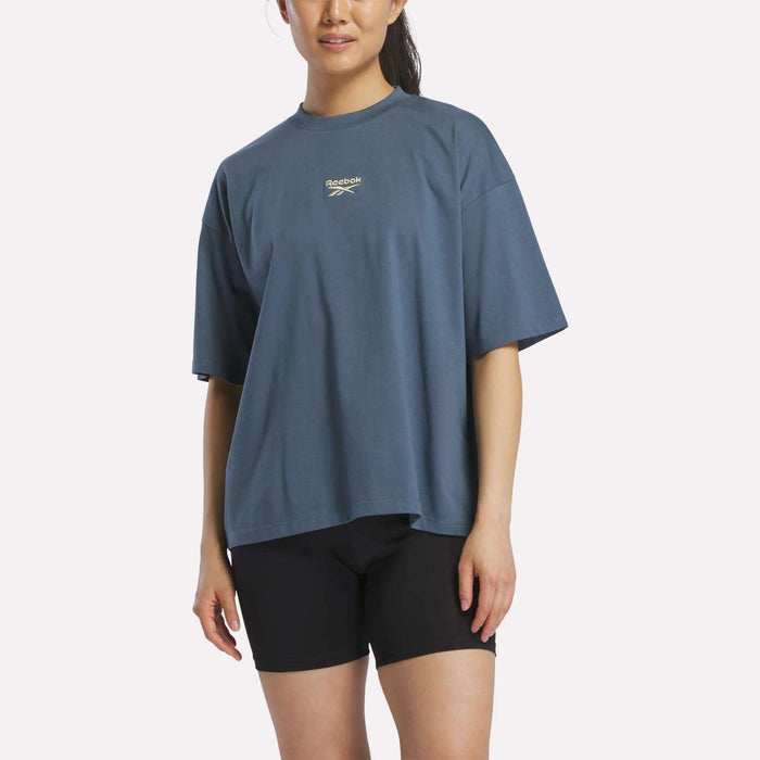 A woman in a loose-fitting Reebok Women's Boxy T-Shirt and black shorts stands against a plain light background.