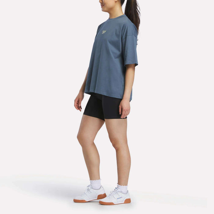 A person stands sideways against a plain white background wearing a Reebok Women's Boxy T-Shirt in soft cotton jersey, paired with black shorts, white socks, and white sneakers with brown soles.