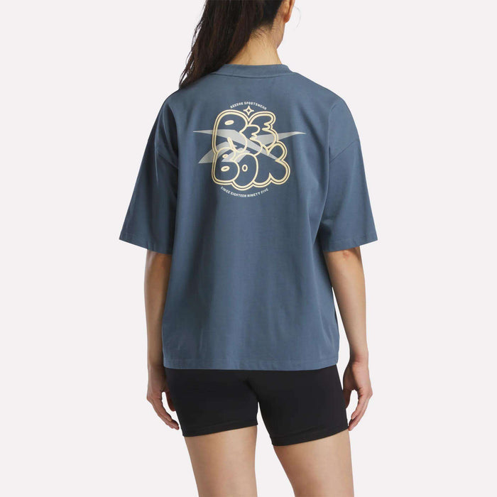 A woman with long dark hair in a ponytail wears a Reebok Women's Boxy T-Shirt made from soft cotton jersey with a large stylized logo on the back and black shorts, standing against a plain light background.