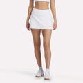 A woman, head not visible, faces forward wearing the Reebok Women's Break Point Skort with a white sports bra, white sneakers, and light blue socks against a plain white background.