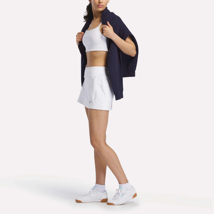 A woman wears a sporty look with a white crop top, Reebok Women's Break Point Skort, white sneakers, and a dark jacket over her shoulders. She stands with her head slightly turned, face partially out of frame.