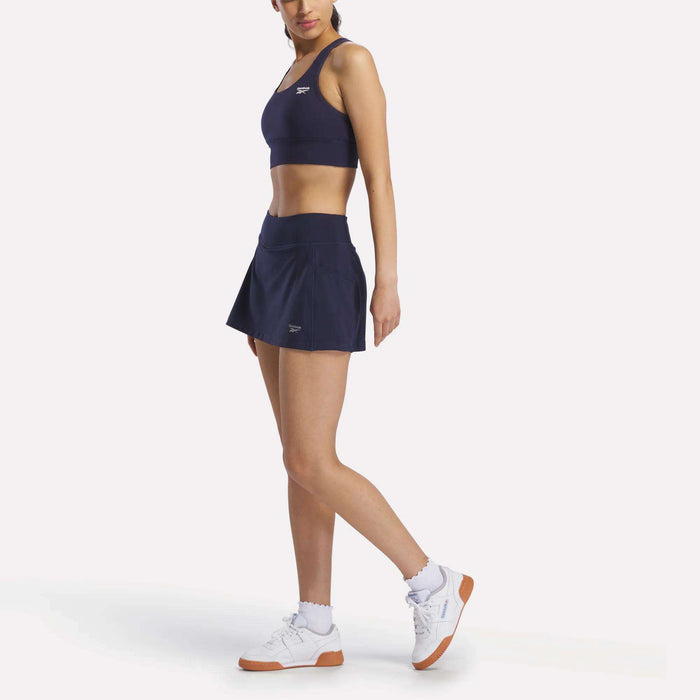 A woman poses against a plain white background wearing the Reebok Women's Break Point Skort in navy blue with a matching sports bra, white socks, and white sneakers with brown soles.