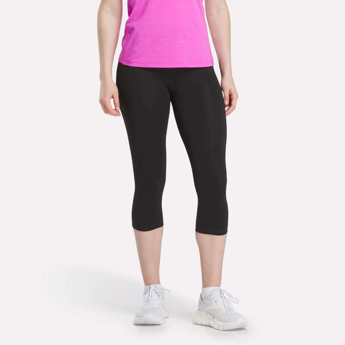 Only the lower half of a person is visible, showing them in bright pink T-shirt, Reebok Women's Capri Leggings, and white sneakers against a plain white background.