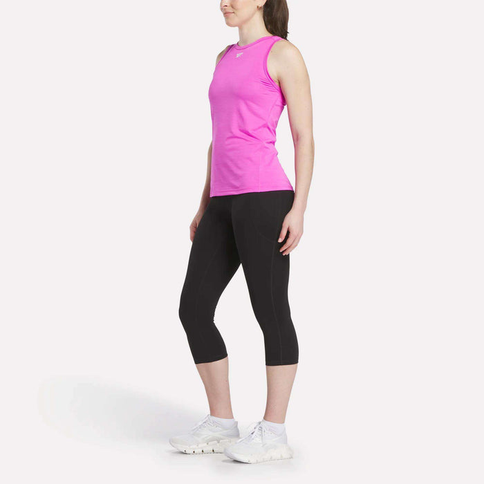 A woman faces slightly to the side, wearing a sleeveless bright pink athletic top, Reebok Women's Capri Leggings, and white sneakers against a plain white background.