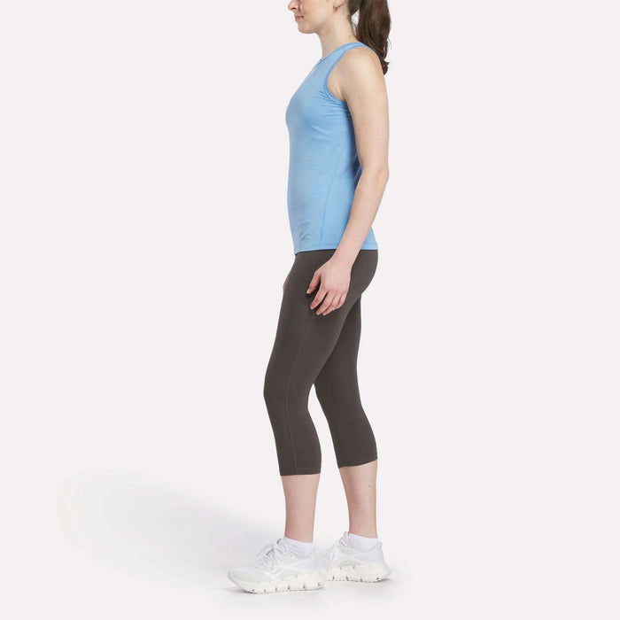 A woman stands in profile against a white background, wearing a blue sleeveless top, white athletic shoes, and dark gray Reebok Women's Capri Leggings.