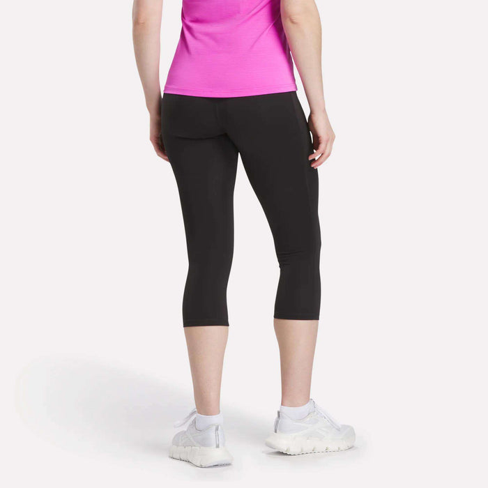 A person stands facing away, hands relaxed by their sides, wearing a bright pink short-sleeve top, Reebok Women's Capri Leggings with an adjustable waistband, and white athletic shoes.
