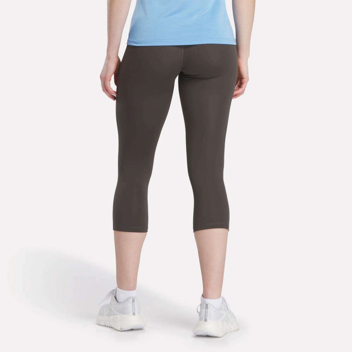 A person is shown from the back, standing on a light background, wearing a light blue shirt, Reebok Women's Capri Leggings in dark gray, and white sneakers.