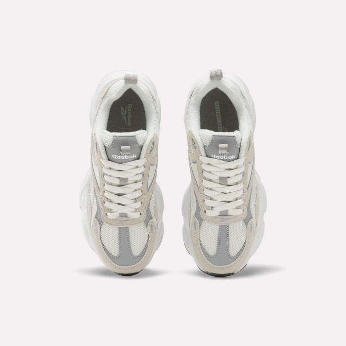 A pair of Reebok Women’s Charge Shoes in white and light gray is viewed from above, displaying the laces and the Reebok logo on each tongue against a plain white background. These women’s athletic shoes feature a subtle chunky design for enhanced style and support.