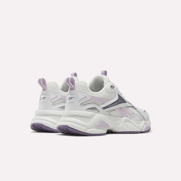 A pair of Reebok Women’s Charge Shoes in white and lavender are displayed from the back and side on a plain light background. Featuring mesh and suede construction with lavender accents and thick soles, these sneakers are perfect for your next workout.