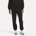 A person wearing Reebok Women’s Cinch Sweatpants in black, paired with a black sweatshirt and white sneakers, stands against a plain light background for a sleek, modern look.