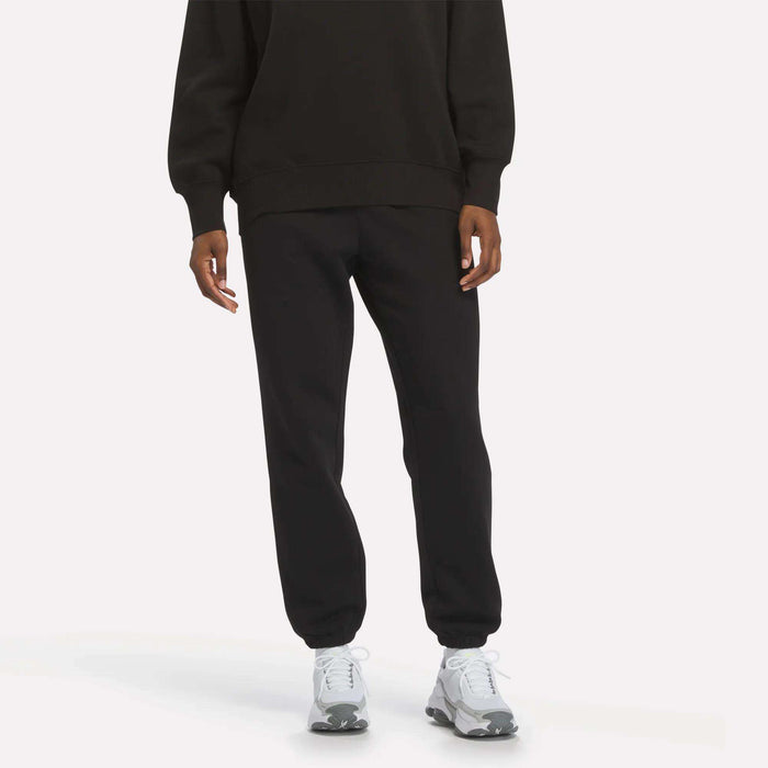 A person wearing Reebok Women’s Cinch Sweatpants in black, paired with a black sweatshirt and white sneakers, stands against a plain light background for a sleek, modern look.