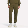 A person wearing Reebok Women’s Cinched Joggers in Olive Stone, a matching sweatshirt, and white sneakers stands against a plain white background.