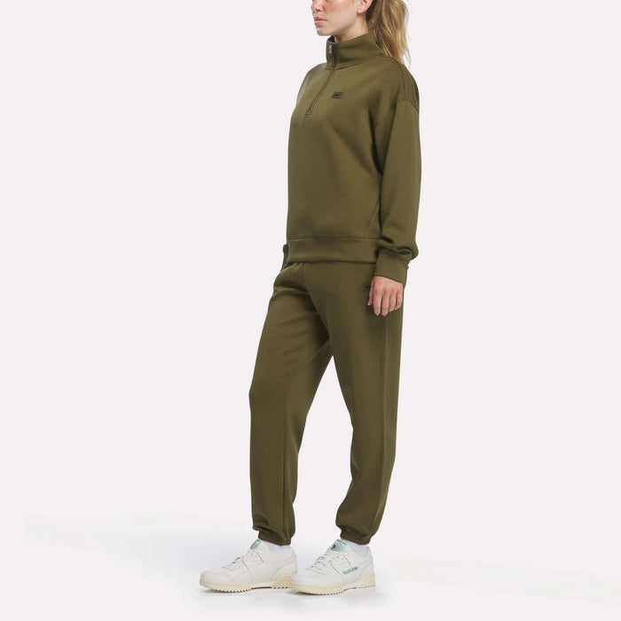 A woman wearing Reebok Women’s Cinched Joggers in olive green and white sneakers stands against a plain white background, gazing off to the side.