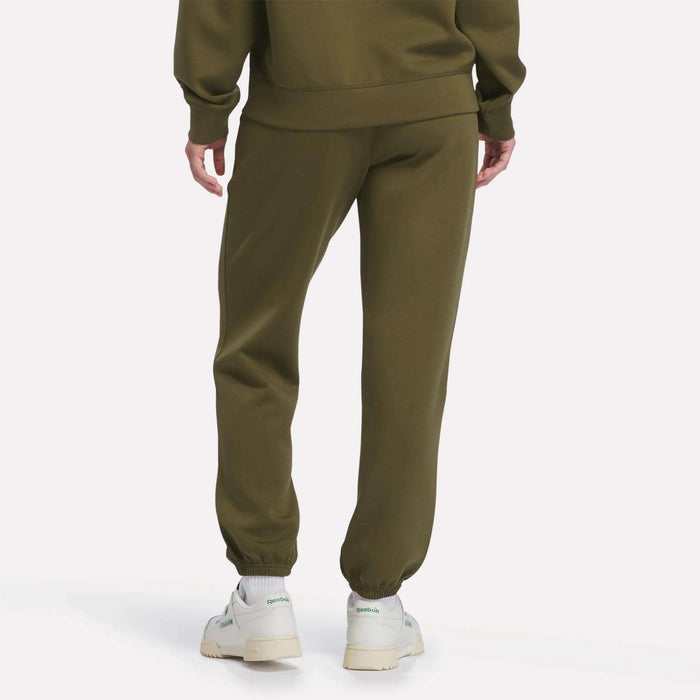 A woman is shown from the back wearing Reebok Women’s Cinched Joggers in olive green with a matching sweatshirt. She completes the look with white sneakers featuring green accents, standing against a light background.