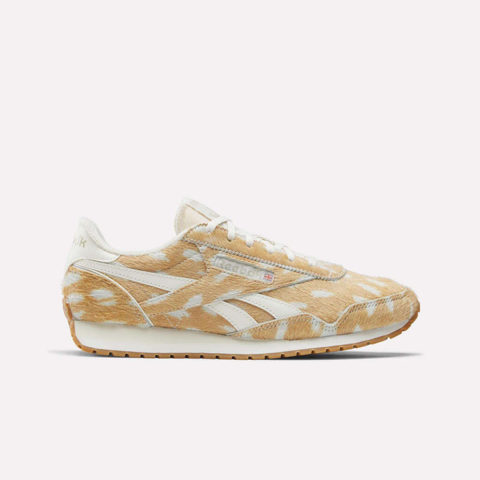 A side view of the Reebok Women's Classic AZ Shoes shows a light tan and white cow-print pattern, white laces, and a gum rubber sole, highlighting the sneakers' retro style against a plain white background.