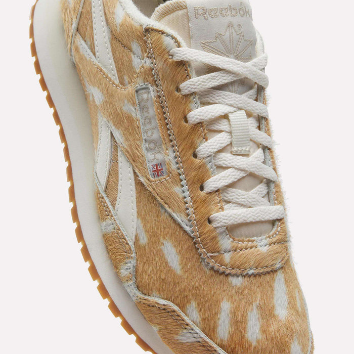 The Women's Classic AZ Shoes by Reebok feature a tan and white spotted faux fur upper, white laces, a gum sole, retro vintage-inspired style, the Reebok logo, and a small Union Jack flag on the side.