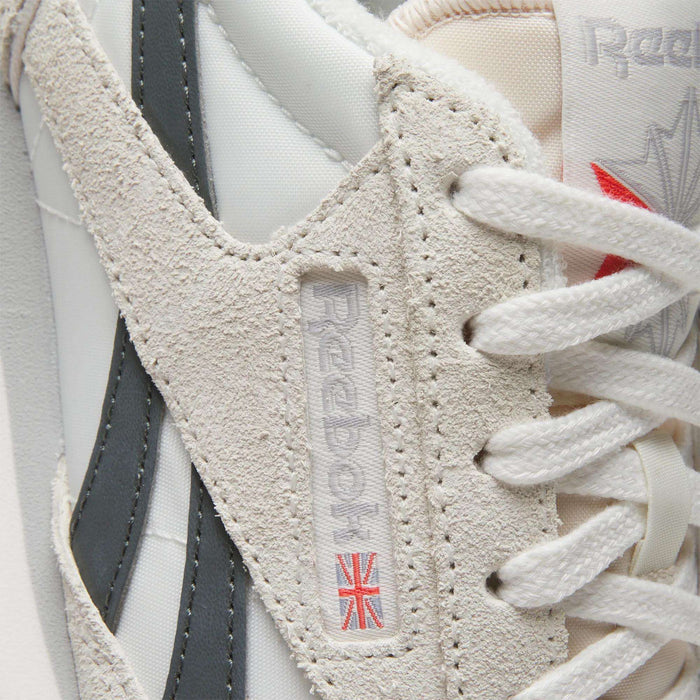 Close-up of the Women's Classic AZ Shoes by Reebok in white and beige, featuring suede and leather accents plus a woven logo tag with a small Union Jack near the laces—ideal for vintage sneaker enthusiasts.