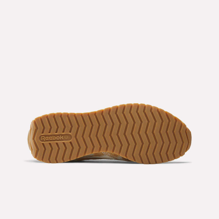 The image shows the bottom view of the Reebok Women's Classic AZ Shoes, featuring a light brown zigzag-patterned rubber sole with the embossed Reebok logo near the heel, showcasing its retro style on a plain white background.