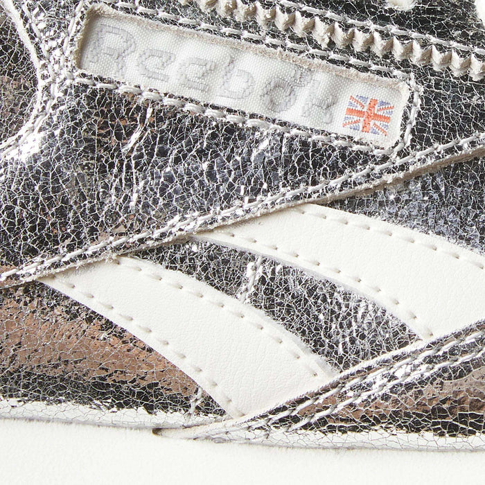 Close-up of a metallic silver Women’s Classic AZ Shoe by Reebok, featuring a textured surface, white stripes, and a label displaying the Reebok logo alongside a small British flag—showcasing the signature details of these iconic retro running shoes.