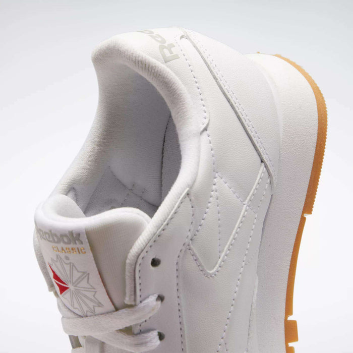 A close-up of the Reebok Women's Classic Leather Shoes in white with a gum sole, subtle stitching, cushioned midsole, Reebok logo, and star emblem on the tongue. The background is plain and light-colored.