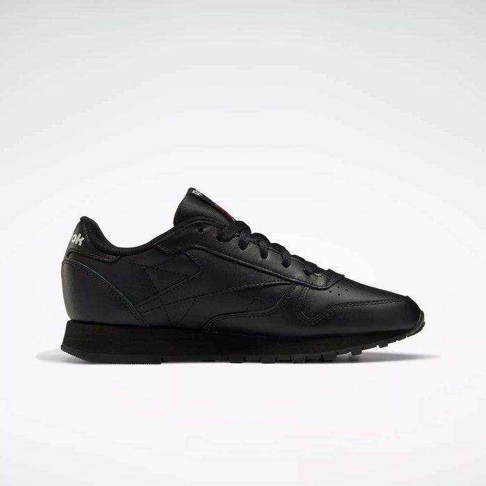 The Reebok Women's Classic Leather Shoes are shown in profile on a white background, featuring a low-top black design, cushioned midsole, black laces, and a textured rubber sole.