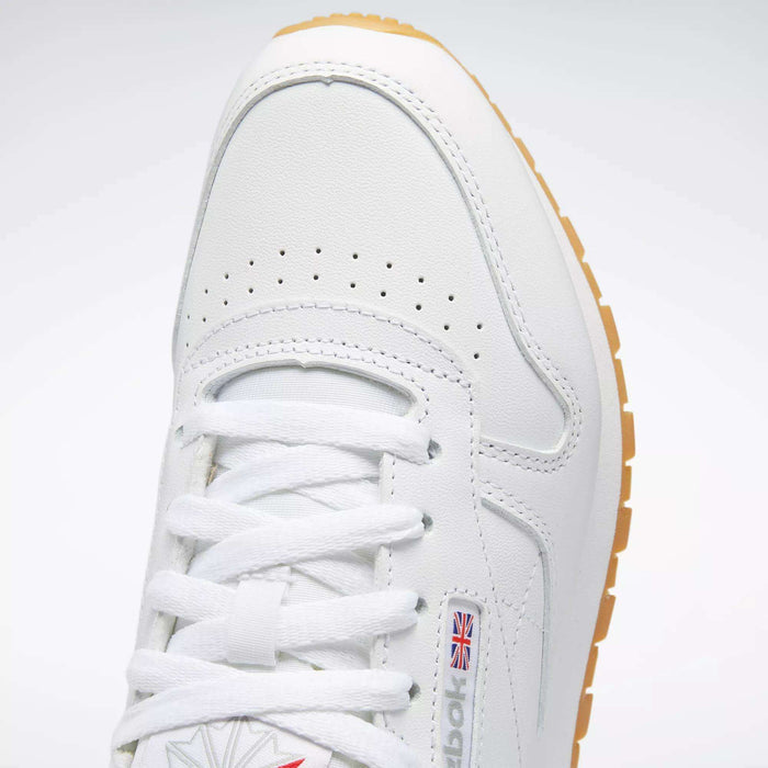 Close-up of Reebok Women's Classic Leather Shoes in white, featuring white laces, perforated toe, cushioned midsole, small British flag logo on the side, and a light brown rubber sole.
