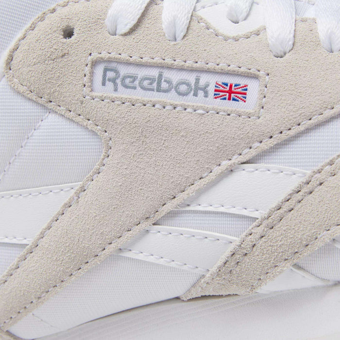 Close-up of Reebok Women's Classic Nylon Shoes in white with beige suede accents, white laces, and the Reebok logo featuring a small UK flag—stylish everyday sneakers.
